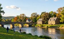 A picturesque view of New Bridge Landing State Park in Bergen County, NJ, capturing its historical significance. In the foreground, a serene river banks with lush green grass and a few visitors casually strolling and admiring the scenery, wearing casual clothing. The middle ground features the historic sandstone bridge and replicas of 18th-century homes, bathed in soft, warm sunlight, reflecting a peaceful afternoon. In the background, gentle hills and vibrant trees create a scenic backdrop. The atmosphere is tranquil and inviting, with a clear blue sky dotted with fluffy white clouds. The image is captured with a wide-angle lens to enhance the depth and immersion. The lighting is natural, highlighting the beauty of the park and its rich history.
